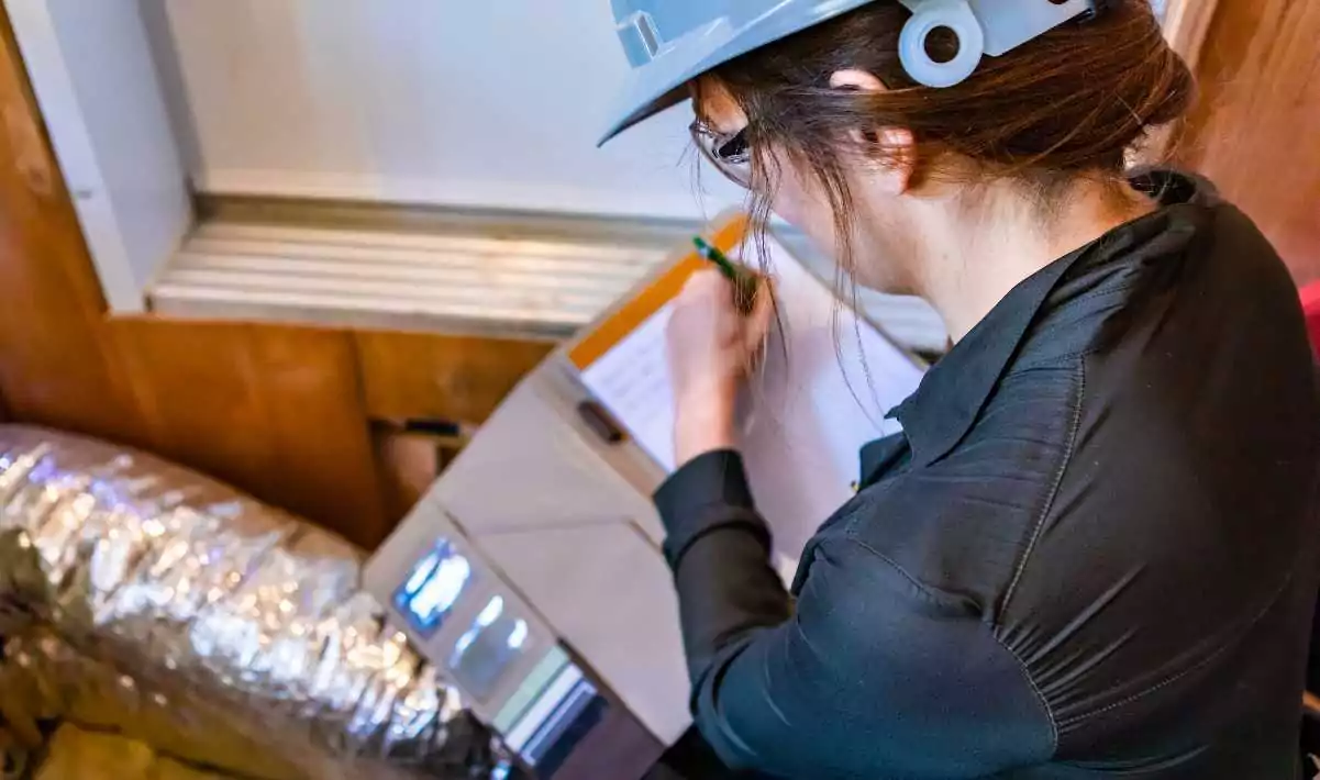 Inspector making notes on a clipboard near a window and exposed ducting.