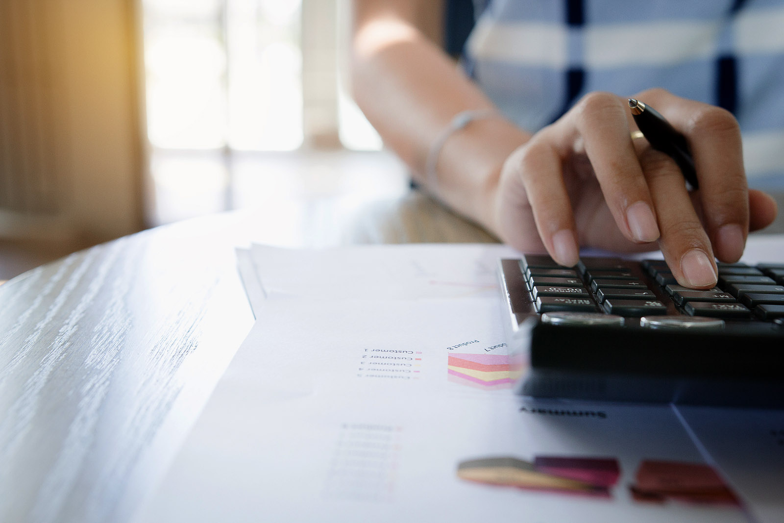 Woman holding pen and using calculator with same hand, at a home desk with financial paperwork.